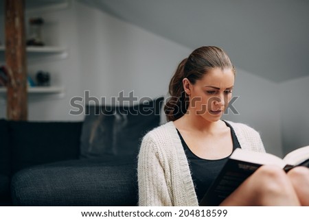 Image of young woman reading a book. Caucasian female at home sitting by a couch with a novel.