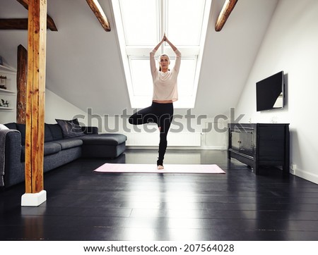 Full length image of fit young woman standing on one leg and meditating in her living room. Caucasian female doing yoga at home.