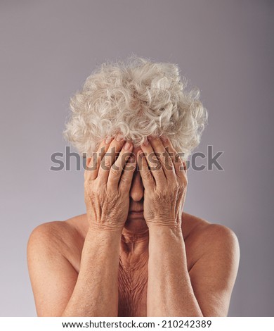 Portrait of  a upset old woman with her head in her hands against grey background. Sad senior woman covering her face.