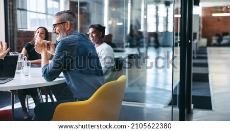 Photo of Cheerful mature businessman attending a meeting with his colleagues in an office. Experienced businessman smiling while sitting with his team in a meeting room. Creative businesspeople working together