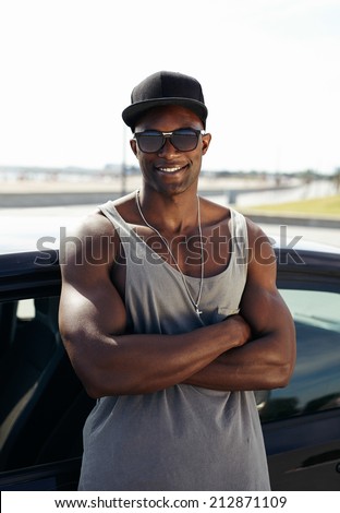 Portrait of handsome african guy with his arms crossed leaning against a car smiling. Stylised muscular guy wearing sunglasses and cap outdoors