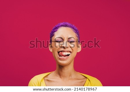 Quirky and vibrant. Excited young woman sticking her tongue out with her eyes closed while standing against a pink background. Fashionable young woman wearing makeup with purple hair in a studio. Foto stock © 