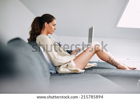 Side view of young lady sitting on sofa using laptop. Caucasian female model on couch surfing internet on laptop computer at home.