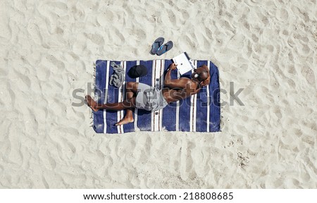 Shirtless young african man lying on a mat reading a book on a sunny day. Young guy relaxing on beach reading and listening to music on headphones.