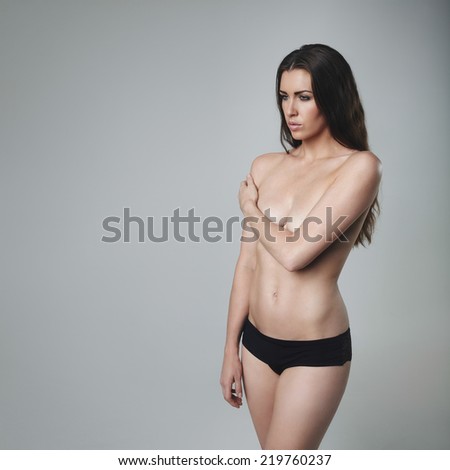 Semi-dressed fashion model standing on grey background looking away at copy space. Brunette woman wearing panties covering her breast with her hand.