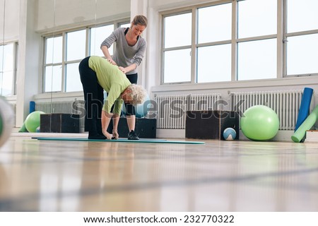 Senior woman bending forward and touching her toes being helped by personal trainer at health club. Elder woman doing back exercise with help from physical therapist at gym.