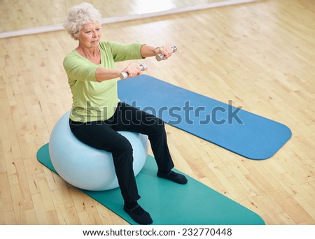 Senior female sitting on a fitness ball and lifting dumbbells. Old woman exercising with weights at gym.