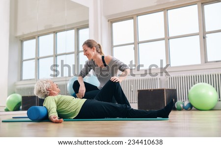 Senior woman having a friendly chat with her personal trainer while exercising at gym. Elder woman doing pilates with gym coach.