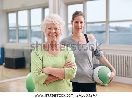 Portrait of happy senior woman standing with her arms crossed and her personal trainer in background gym. Caucasian elderly woman at health club with gym instructor.