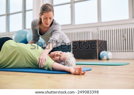 Senior women lying on exercise mat doing stretching workout for back muscles with coach assistance. Female trainer helping elder woman in stretching.