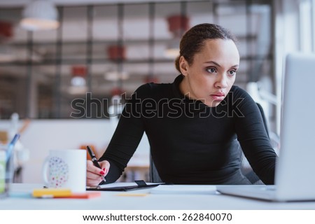 Image of young businesswoman looking at laptop while working at her desk. Female web designer taking notes from internet.