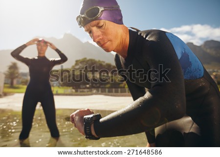 Focused young man checking his watch while in wetsuit at the lake. Man looking at watch after practice run. Triathletes getting ready for race.