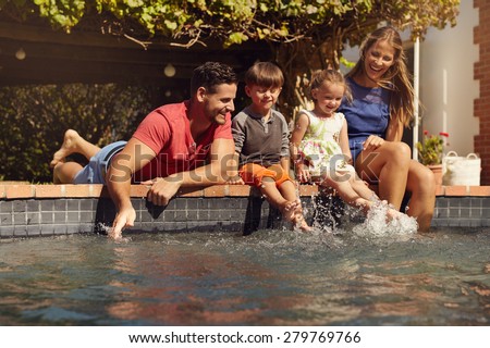 Caucasian family having fun by their swimming pool. Happy young family splashing water with hands and legs while sitting on edge of swimming pool. Kids with parents playing outdoors.