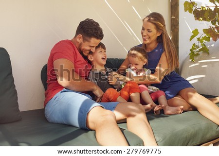 Shot of happy family of four in their backyard having fun, sitting on couch playing. Parents playing with kids laughing in patio.