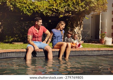 Happy young couple sitting on the edge of swimming pool with their kids enjoying a hot summer day near pool. Couple\'s feet in water and kids playing by outdoors.