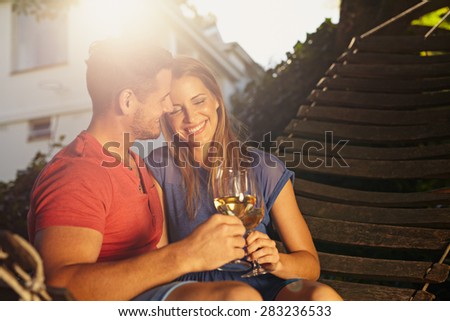 Beautiful young couple toasting wine outdoors. They are sitting on a hammock smiling and drinking wine with bright sunlight in backyard. Young man and woman spending quality time together on weekend.