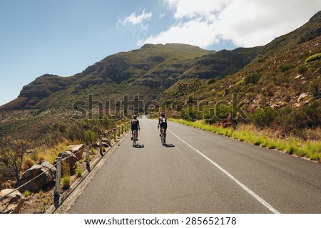 Rear view of two cyclist riding down the country road through mountains. Cycling event of triathlon competition. Triathletes practicing cycling on open country road.