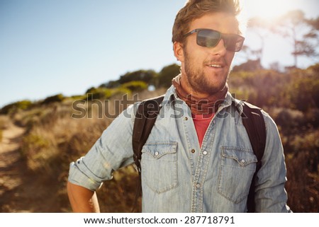 Portrait of happy young man hiking in countryside. Caucasian male model with backpack hiking on sunny day. Summer vacation in countryside.