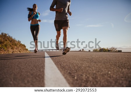 Low angle shot of young woman running on road with man in front on a summer morning. Runners training on country road.