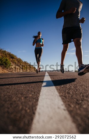 Low angle shot of young woman running on road with man in front. Runners training on country road on summer morning.