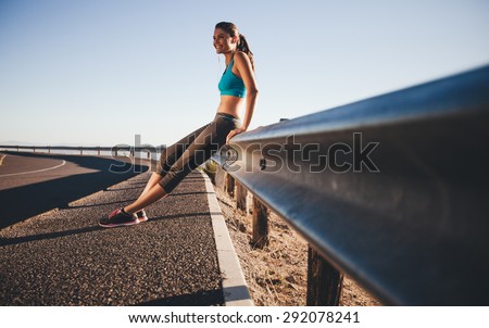 Outdoor shot of happy young woman sitting on highway guardrail after a morning run. Fit woman on country road taking a break after outdoor workout.