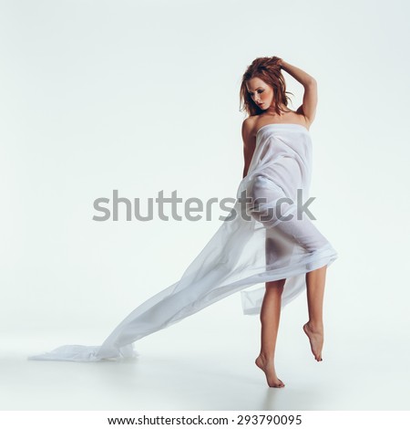 Studio shot of nude brunette posing with a sheer fabric on her body. Full length portrait of caucasian female model naked, wrapped in a transparent cloth posing sensually on white background.