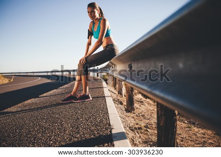 Outdoor shot of young female runner taking a break. Tired young woman sitting on highway guardrail by a country road, resting after running workout.