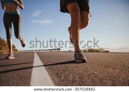 Cropped shot of a young athletes running outdoors. Runners running on a country road on summer morning. Man and woman jogging on road, focus on legs.