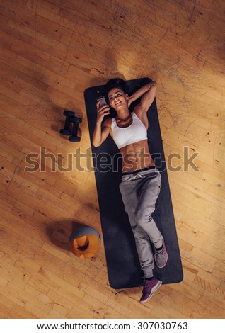 Top view of woman relaxing on yoga mat using mobile phone. Fitness female taking a break lying on exercise mat at gym reading text message on her smart phone.