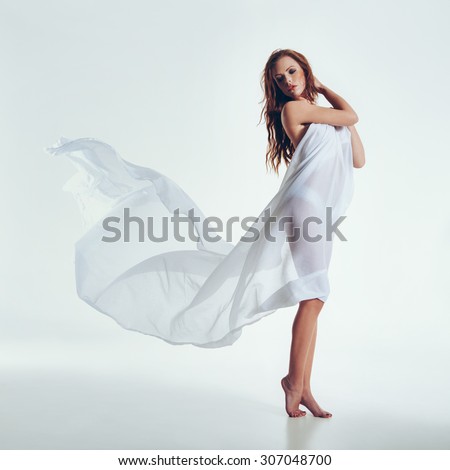 Portrait of a sexy woman with flying textile in the studio. Beautiful young woman wrapped in transparent fabric posing on white background.