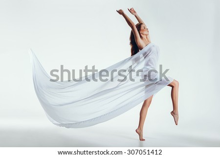 Portrait of a sensual woman with flying textile in the studio. Attractive young woman wrapped in transparent fabric standing on one leg and hands raised over white background.