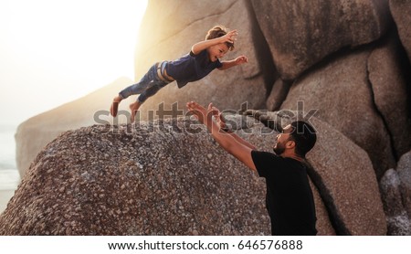 Shot of cute little boy leaping into his fathers arms from a big rock. Father and son enjoying summer holidays at the beach. Foto d'archivio © 