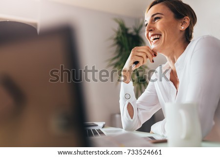 Photo of Smiling woman sitting at her desk in office. Happy business woman sitting in office with fingers touching her chin.