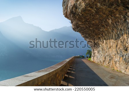 Road along the edge of the cliffs along the lake. Mountain stones hanging from above and secure with a grid. Rays of the sun creates volumetric perspective