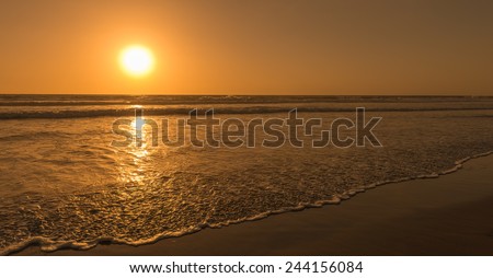 Sun sets over horizon of ocean. Surf on sandy beach. Sky, water and shore in the evening painted in orange, gold color.