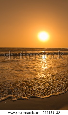 Sun sets over horizon of ocean. Surf on sandy beach. Sky, water and shore in the evening painted in orange, gold color.