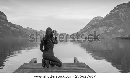 The female figure  is sitting with a camera in hand on a wooden pier. Mountain landscape at sunset, reflected in the lake. Black and white.