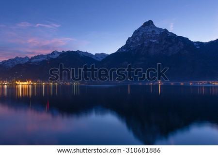 Small town in Swiss Alps. Night sky and clouds sunset painted in red.  Lanterns and lights reflected in water of lake.