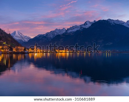 Small town in Swiss Alps. Night sky and clouds sunset painted in red.  Lanterns and lights reflected in water of lake.