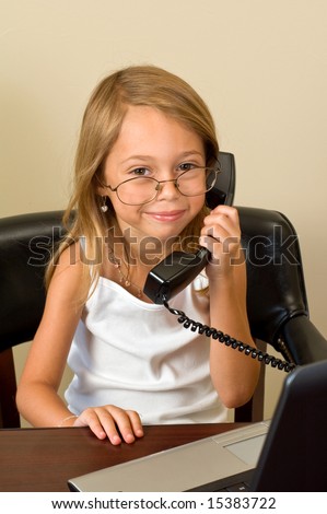 A young girl about 6 pretends to be busy at her notebook computer while wearing glasses too big for her head, talking on a telephone.
