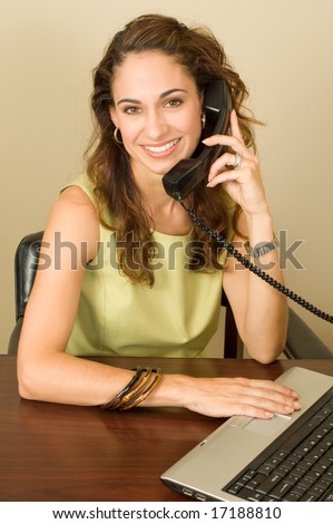 A pretty brunette business professional wearing a green outfit using a notebook computer while on the phone.