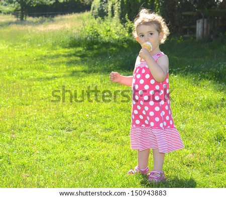 Young girl eating ice cream. Pretty child, toddler, wearing pink and white dress. She has blonde curly hair. Background is green grass.  Bright and colourful.