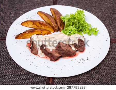 Delicious beef steak with roasted potatoes and fresh lettuce covered with white sauce. Plate located on a brown tablecloth background.