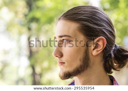 Portrait of handsome young long hair and bearded brunette man at outdoors. Side view. Photo taked against park trees.
