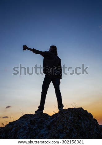 Silhouette of male model taking selfie on the rocks against sunset blue and yellow sky.