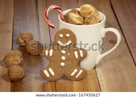 Mug of hot chocolate topped with toasted marshmallows and a candy cane with single gingerbread cookie leaning against mug and walnuts as an accent on wooden table.