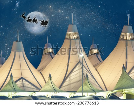 Santa and his reindeers flying over Denver International Airport during the evening.  The tent peaked roofs of the airport are illuminated against an evening sky.