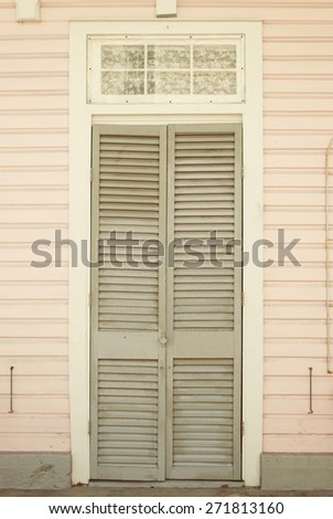 Soft pink-colored house with closed gray shutter door and a  small window above dressed with lace curtains