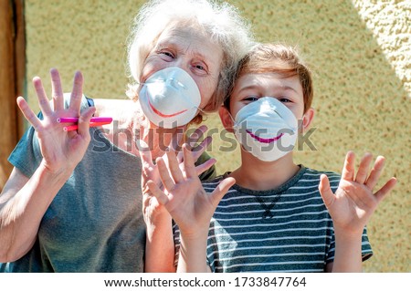 Happy laughing grandmother with grandchild in a respiratory masks plays together near house. Family fun. Stay at home. Drawing a smile on protective masks. Quarantin, isolated. Coronavirus covid-19.