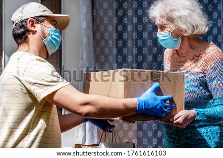 Young male volunteer in mask gives an elderly woman boxes with food near her house. Son helps a single elderly mother. Family support, caring. Quarantined, isolated. Coronavirus covid-19. Donation